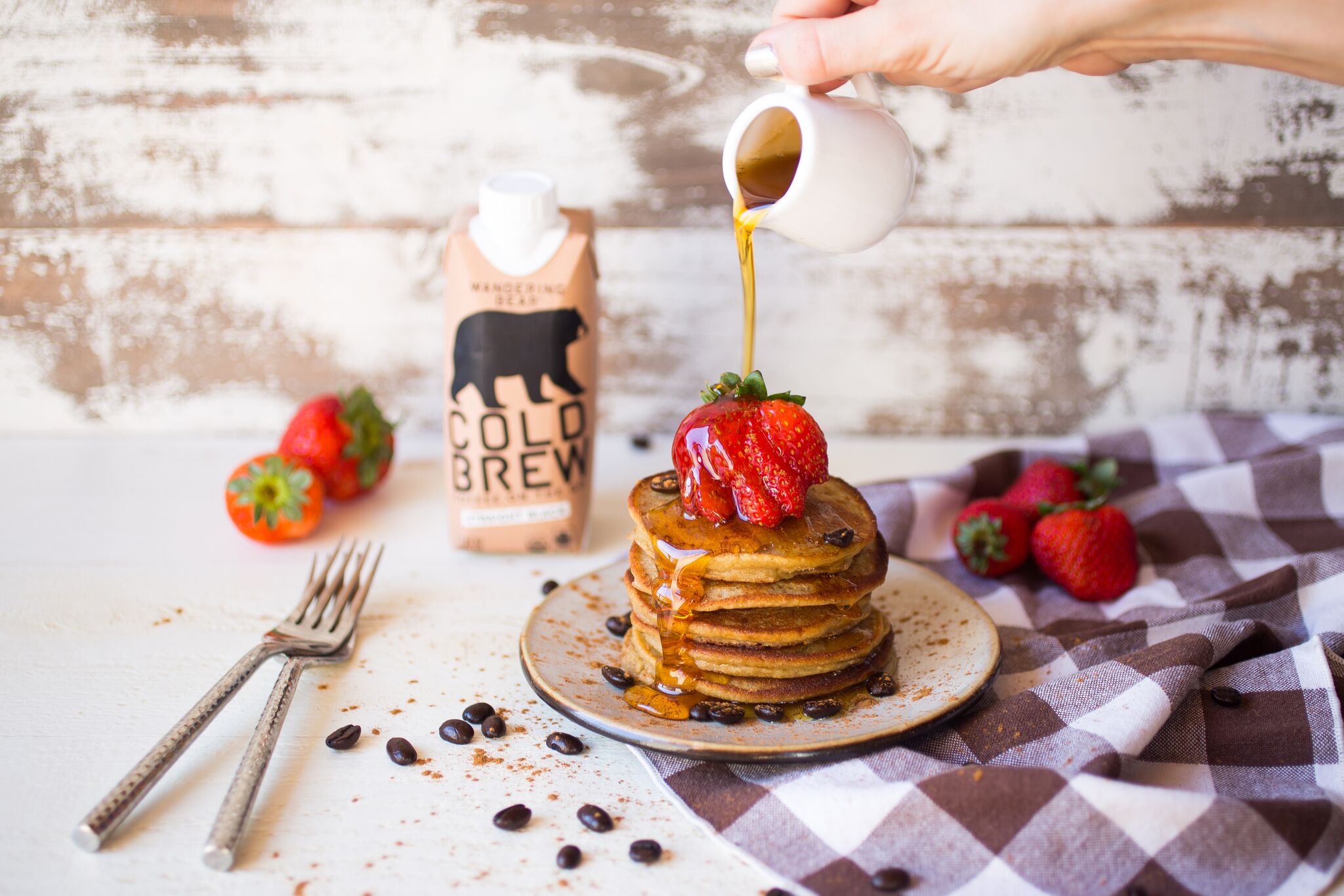 Stack of coffee pancakes topped with a strawberry as syrup is poured, Wandering Bear cold brew carton and strawberries on a rustic wooden table