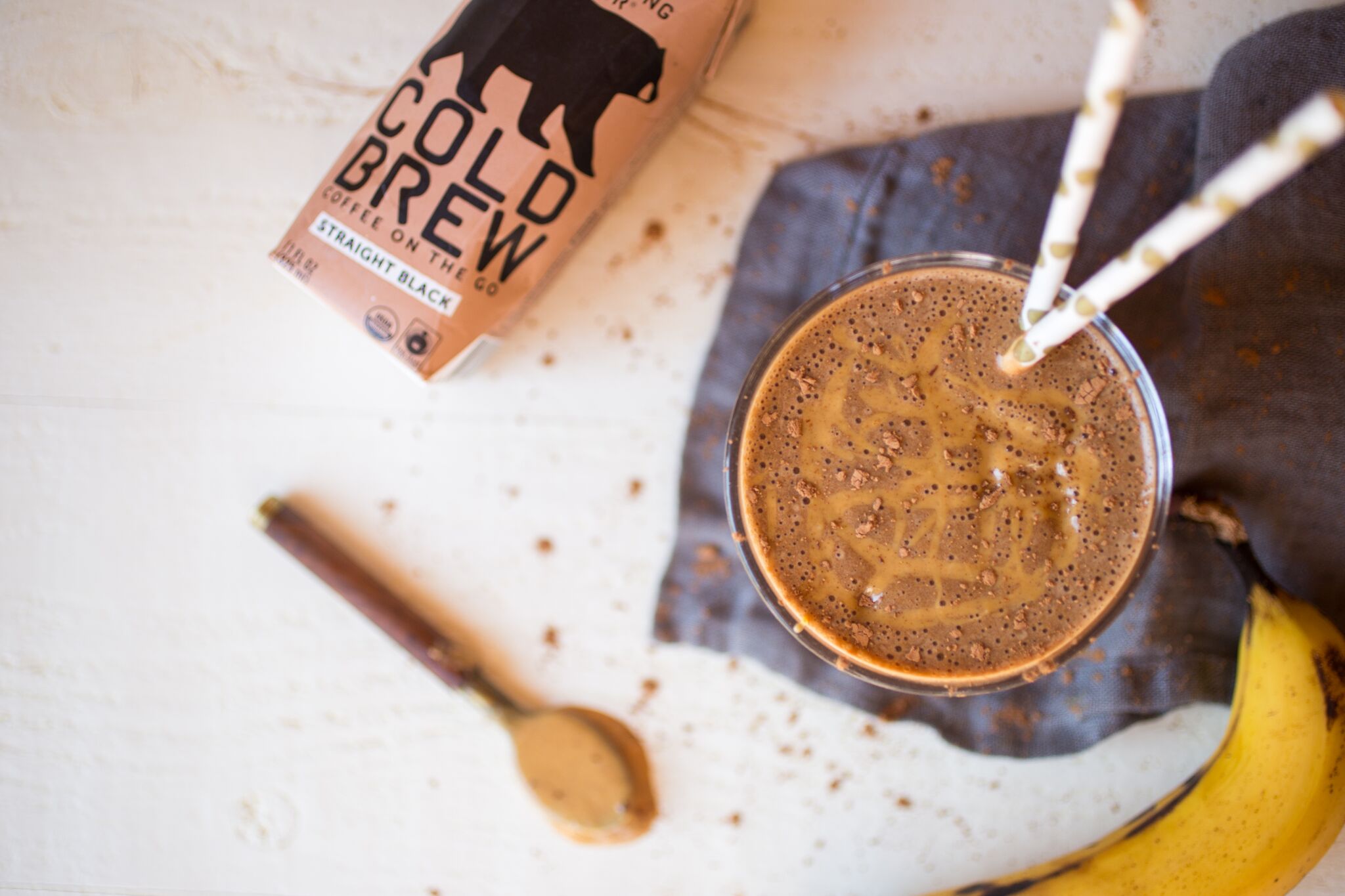 Top view of coffee peanut butter banana smoothie with peanut butter drizzle and chocolate crumbs beside cold brew and banana