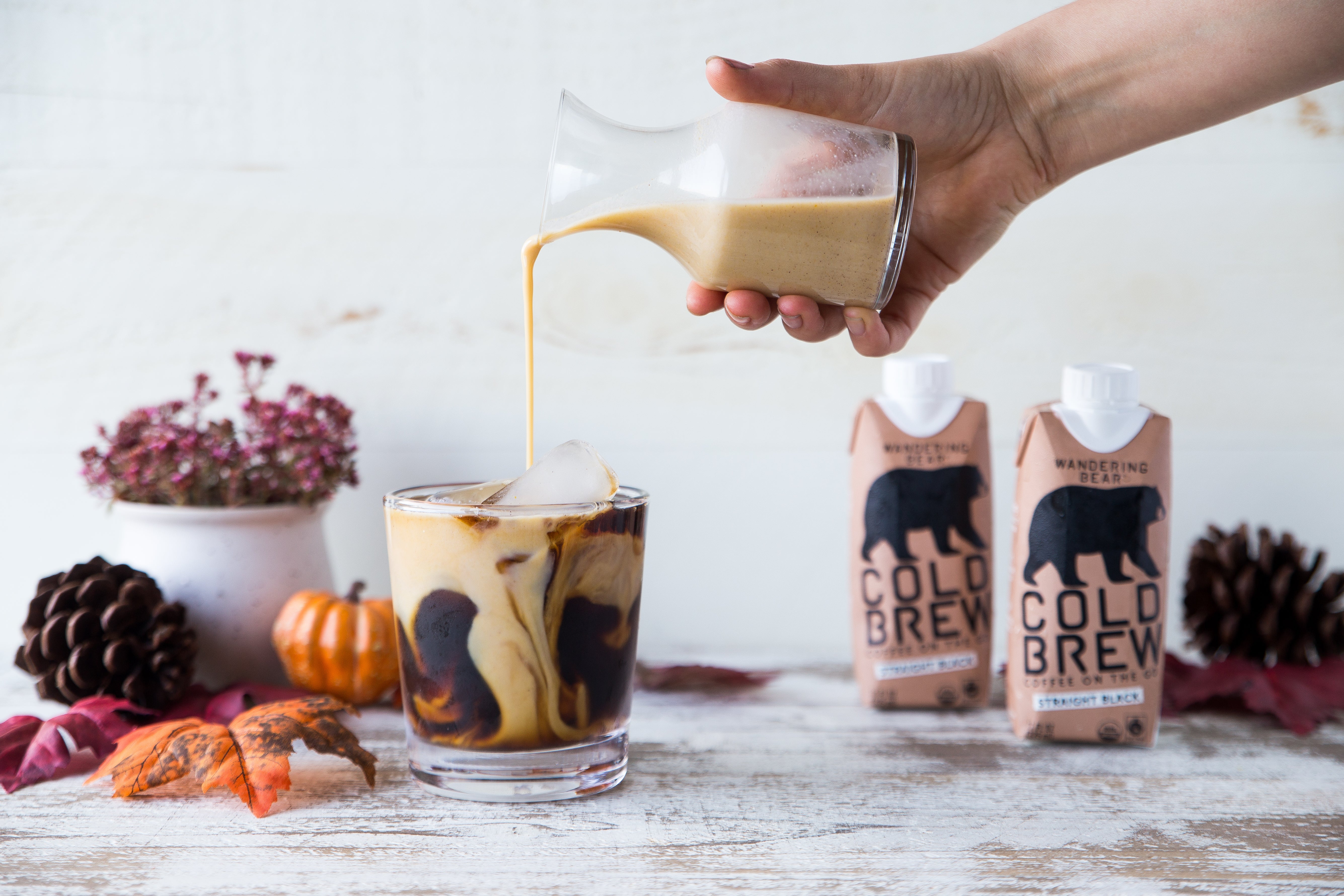 Hand pouring pumpkin-spiced creamer into a glass of iced cold brew with two Wandering Bear cartons and autumn accents