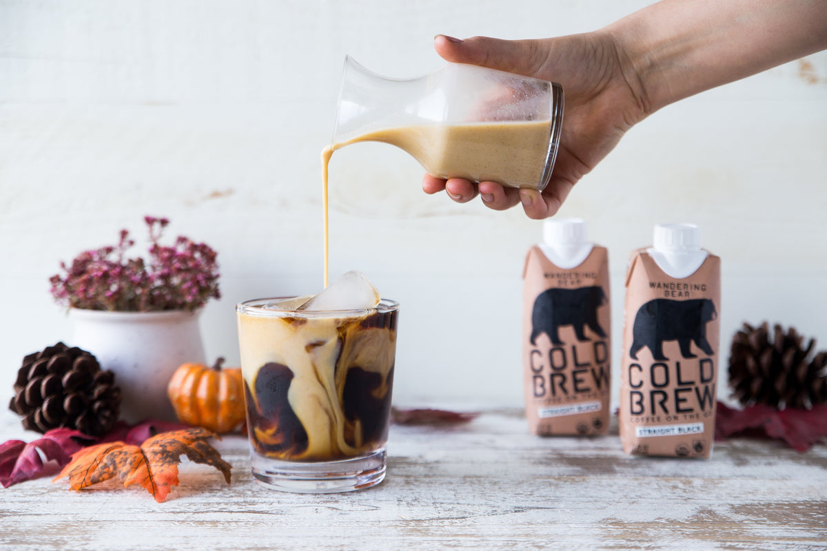 Hand pouring pumpkin spice creamer into a glass of iced cold brew, with Wandering Bear cartons and fall decor