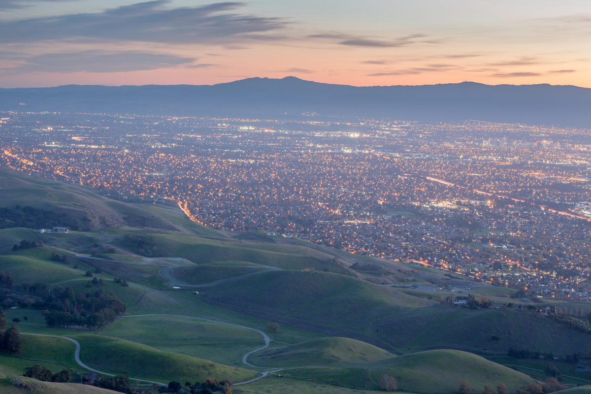 San Jose skyline glowing at dusk above rolling green hills with city lights below