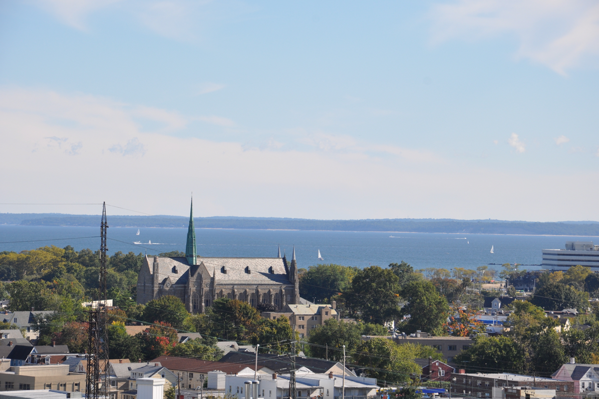 Stone church with green spire above city rooftops, sailboats on the harbor and distant shoreline