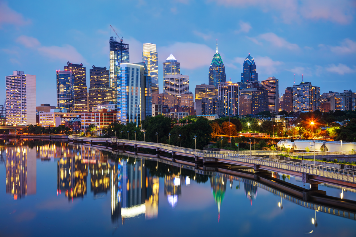 Philadelphia skyline at dusk reflected in the Schuylkill River with lit skyscrapers and a riverside pedestrian path