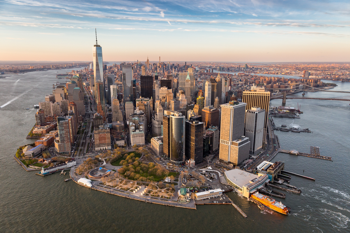 Aerial view of Lower Manhattan skyline at golden hour showing One World Trade Center, waterfront parks, and Staten Island Ferry terminal
