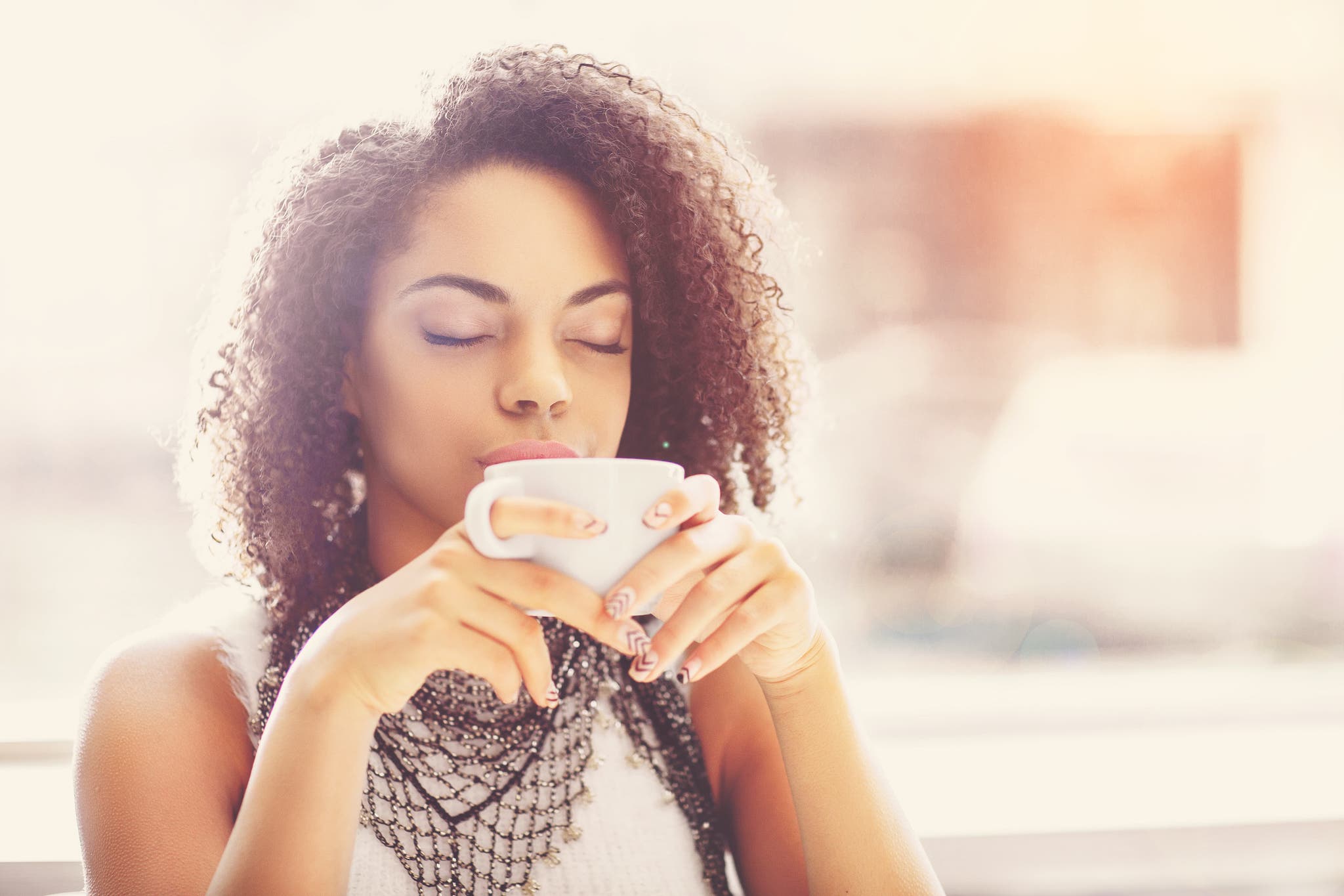 Woman with curly hair holding a white mug, eyes closed and savoring her coffee in warm backlit light