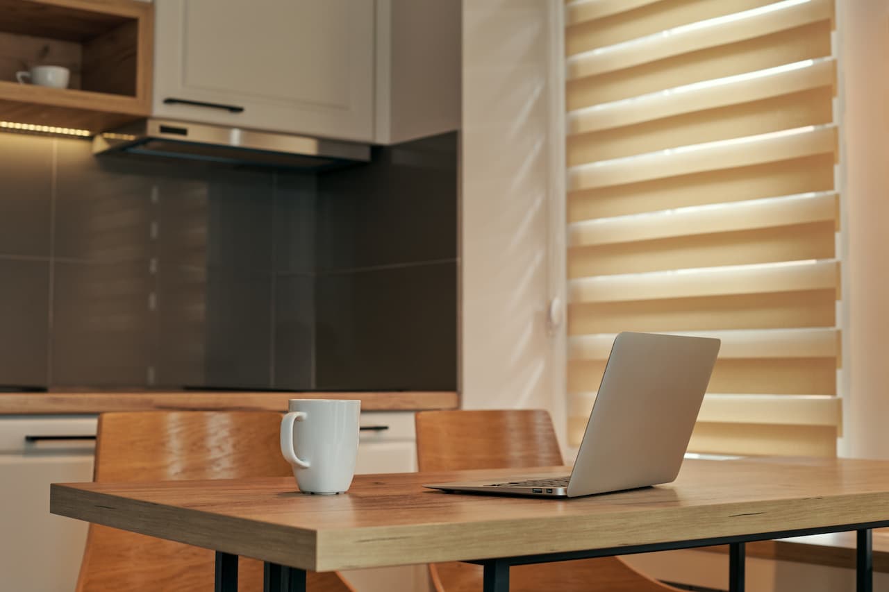 White coffee mug beside an open laptop on a wooden table in a sunlit kitchen with beige blinds