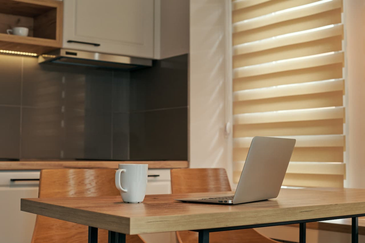 White coffee mug beside an open laptop on a wooden table in a sunlit kitchen with beige blinds