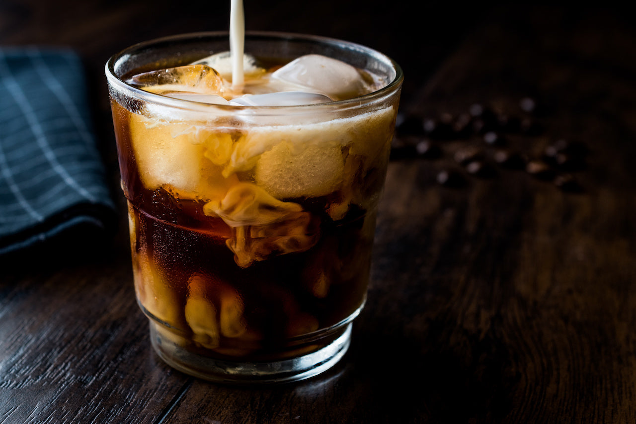 Glass of cold brew with ice as cream is poured in, dark wooden table with scattered coffee beans in the background