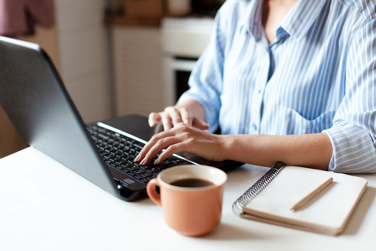 Person typing on a laptop at a home desk with a coffee mug and a notebook nearby