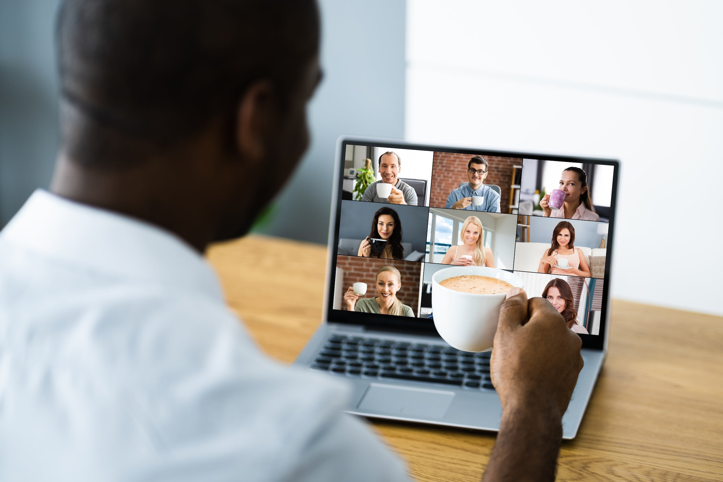 Man holding a coffee mug while joining a virtual coffee break with coworkers shown in a video-call grid on a laptop