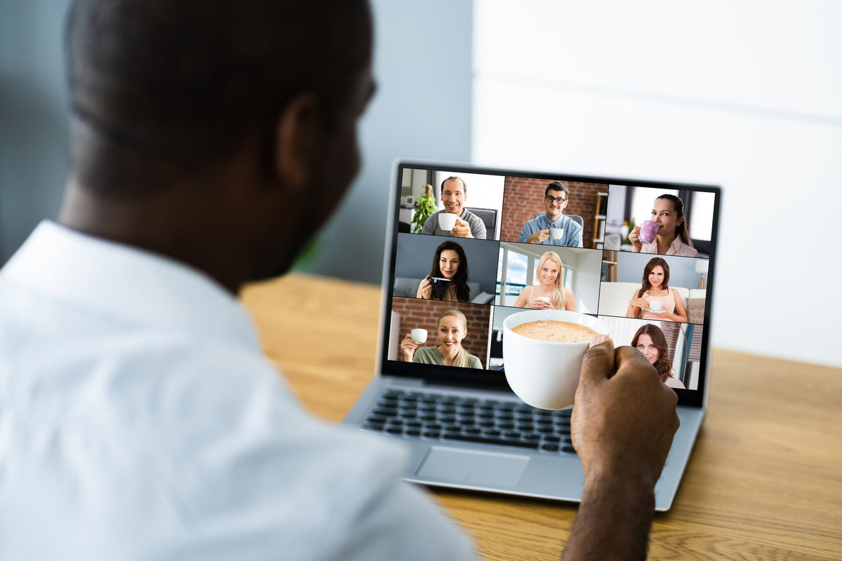 Man holding a coffee mug while joining a virtual coffee break with coworkers shown in a video-call grid on a laptop