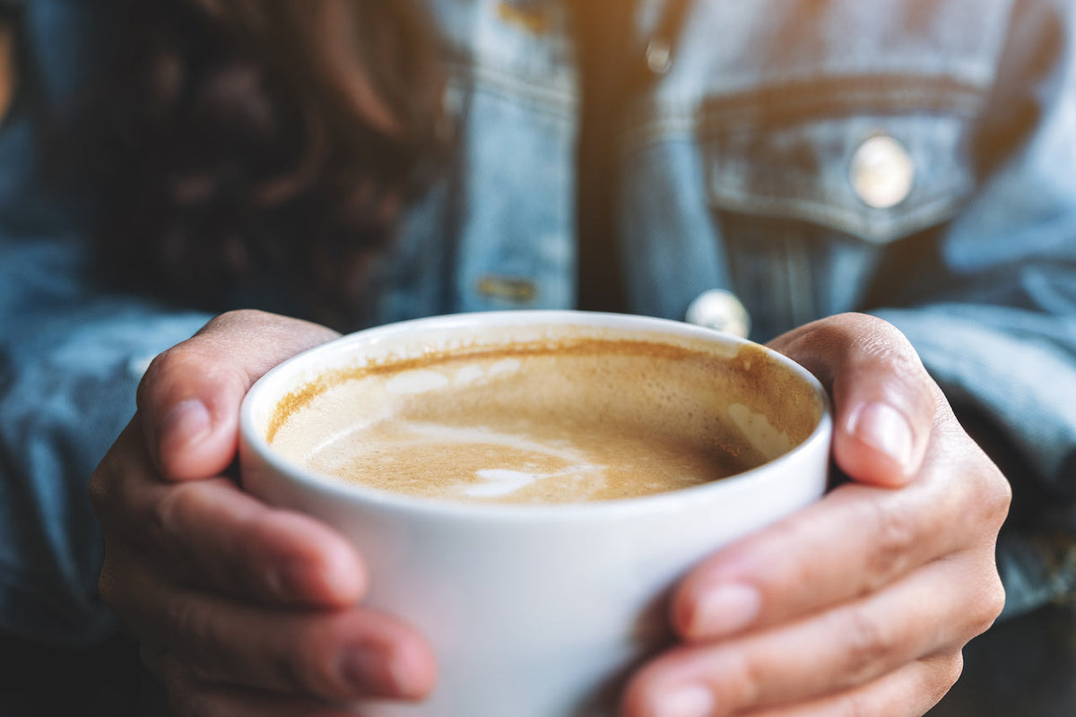 Hands holding a white cup of latte with light foam, person wearing a denim jacket in a soft-lit café setting