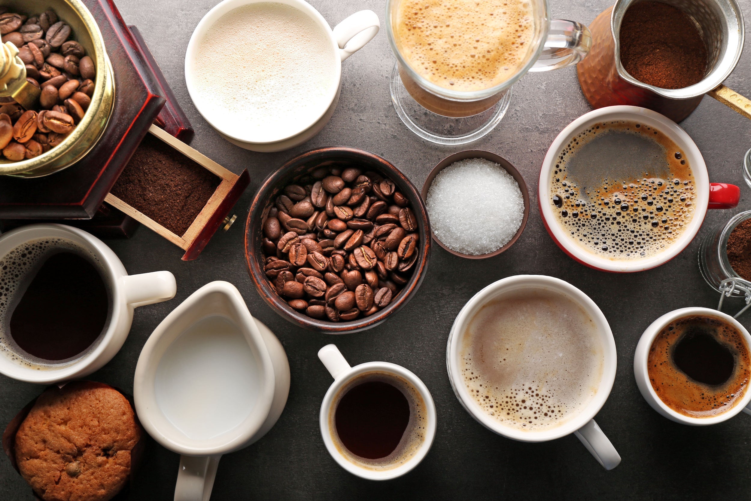 Overhead view of assorted brewed coffees, espresso shots, steamed milk, coffee beans, grinder, and sugar on a dark tabletop
