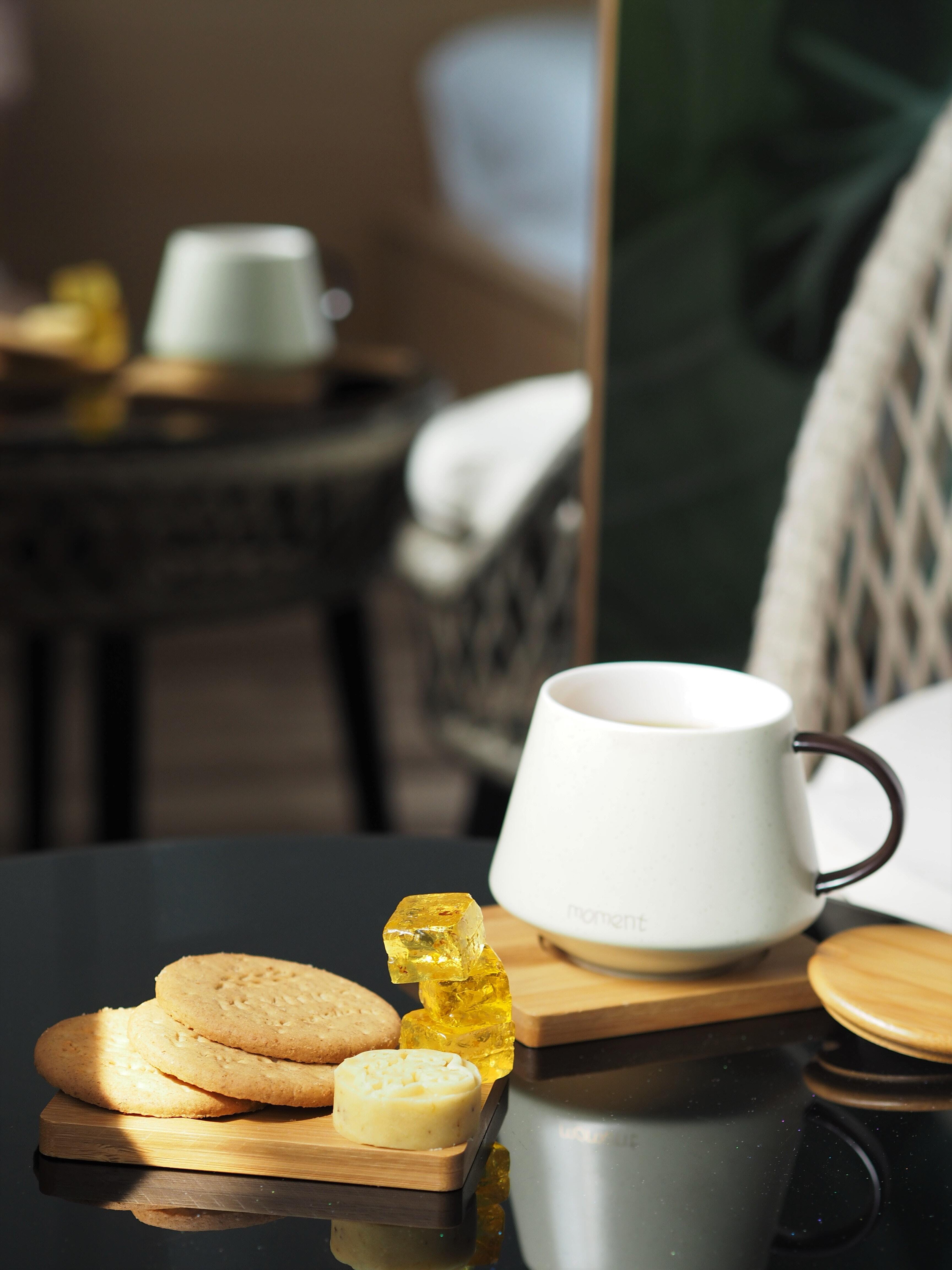 Close-up of a white ceramic coffee mug and stacked biscuits on a wooden board on a dark reflective table with a blurred background
