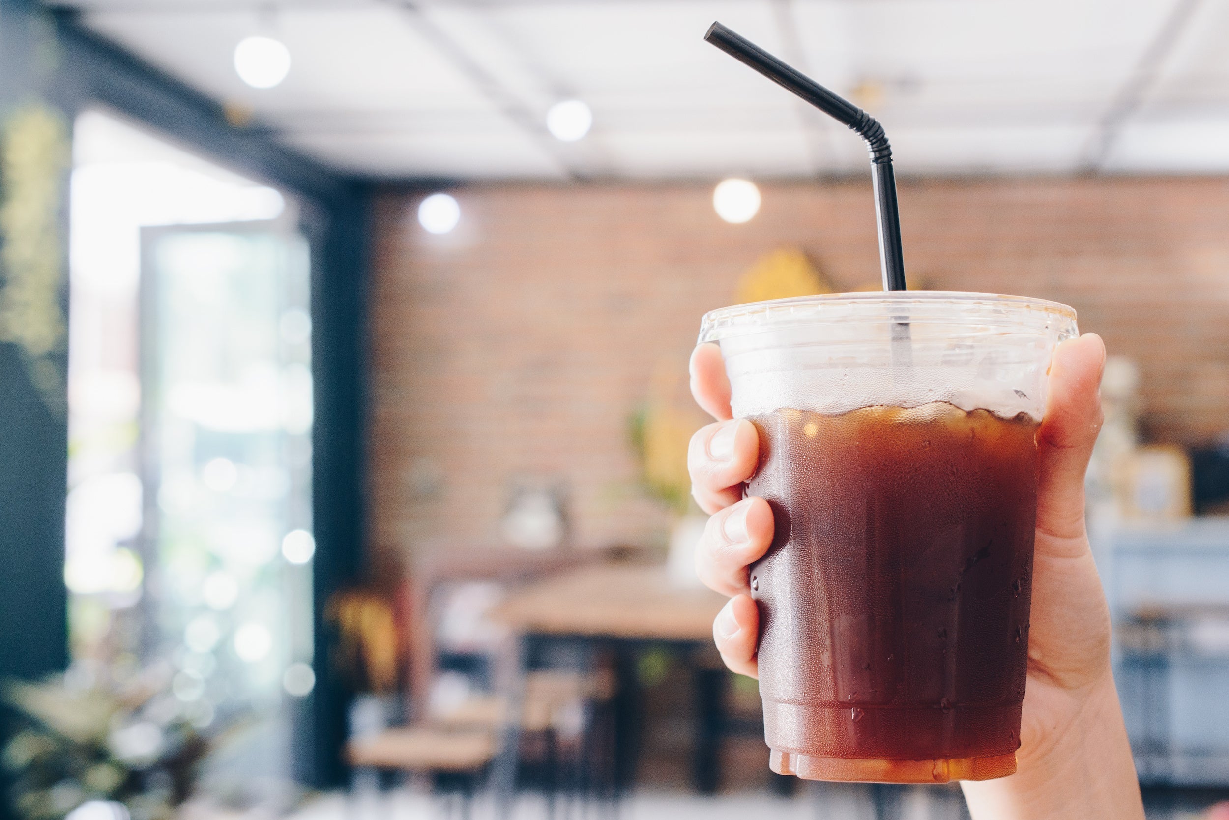 Hand holding a plastic cup of iced cold brew with a black straw in a bright blurred cafe interior