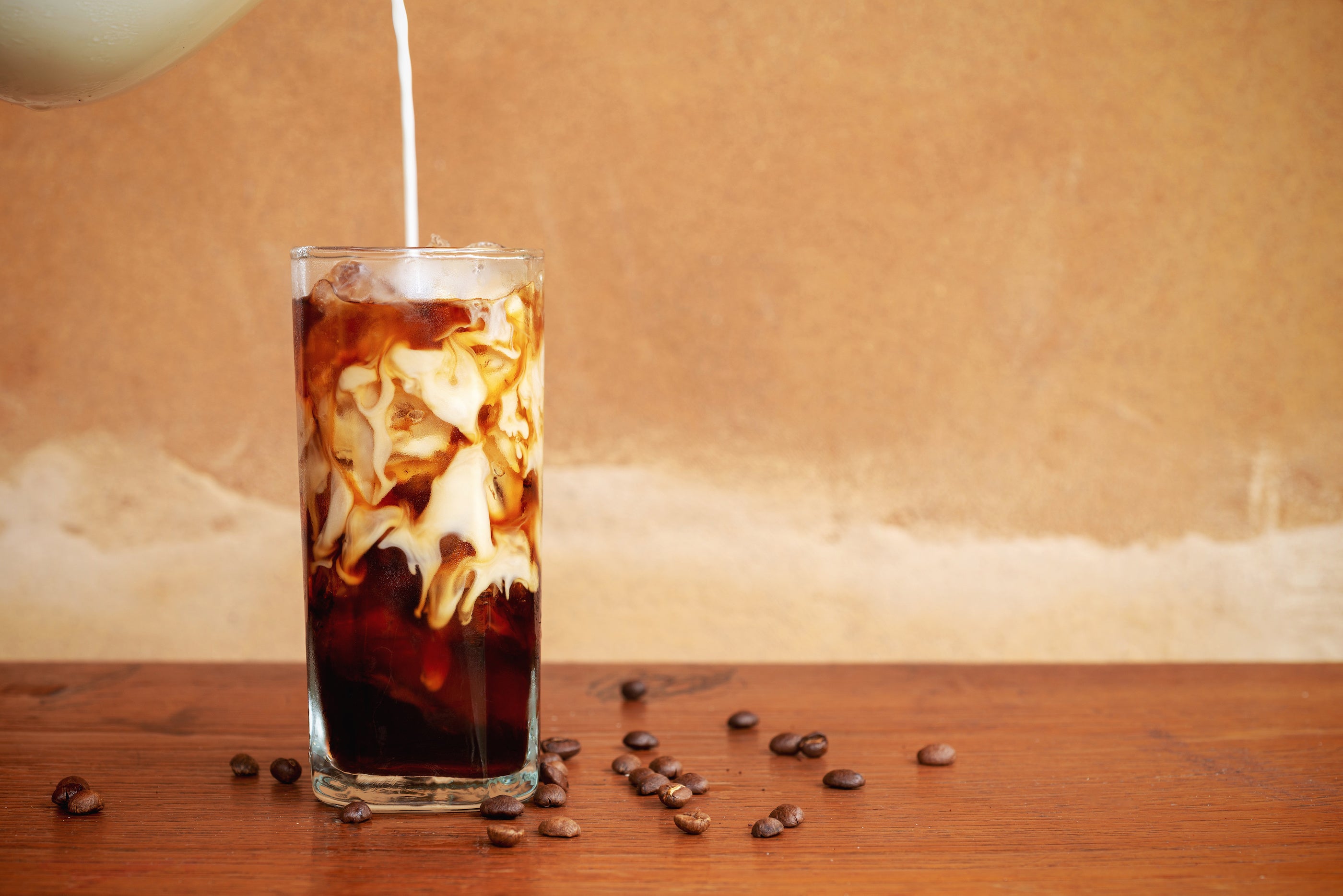 Glass of iced cold brew with cream being poured, coffee beans scattered on a wooden table and warm beige background