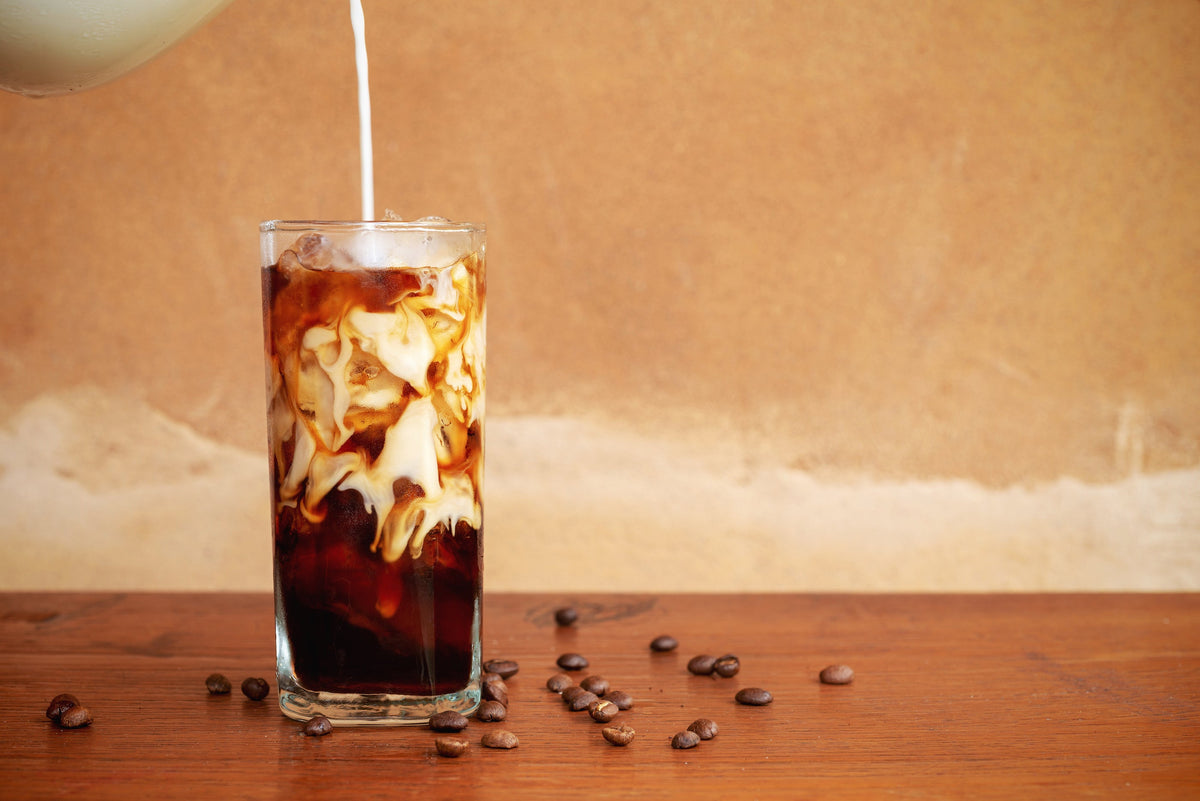 Glass of iced cold brew with cream being poured, coffee beans scattered on a wooden table and warm beige background