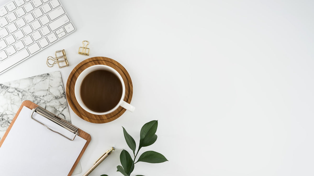 Top-down view of a cup of coffee on a wooden coaster at a tidy office desk with a keyboard, clipboard, pen, and plant
