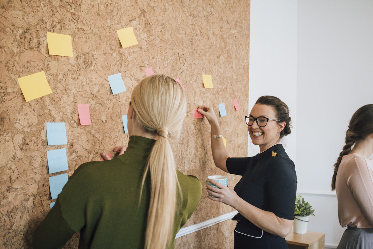 Three coworkers arranging colorful sticky notes on a corkboard while one woman smiles and holds a coffee cup in an office