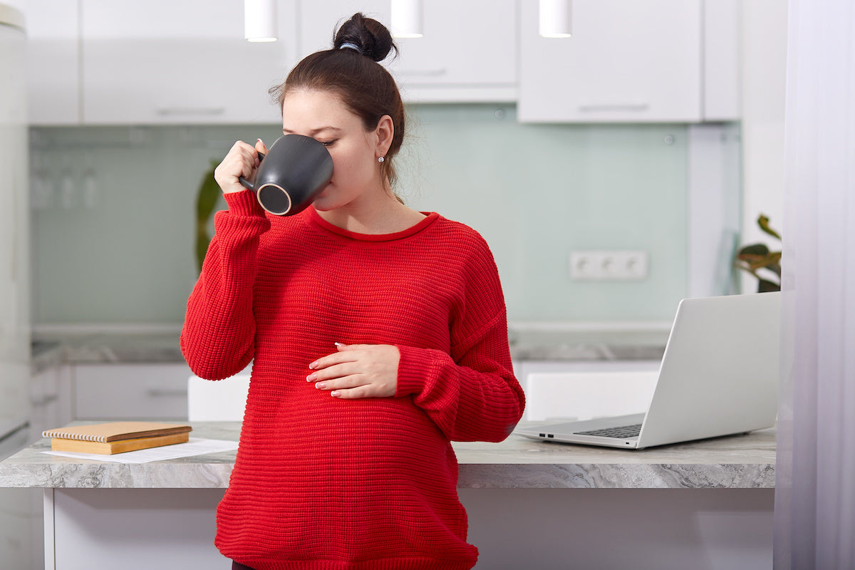 Pregnant woman in a red sweater drinking coffee in a kitchen with one hand on her belly and a laptop on the counter