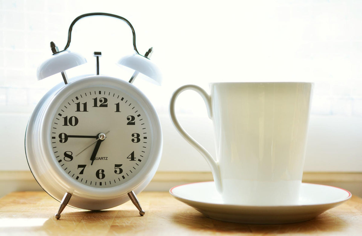 White alarm clock next to a white coffee cup on a saucer on a wooden table by a sunlit window