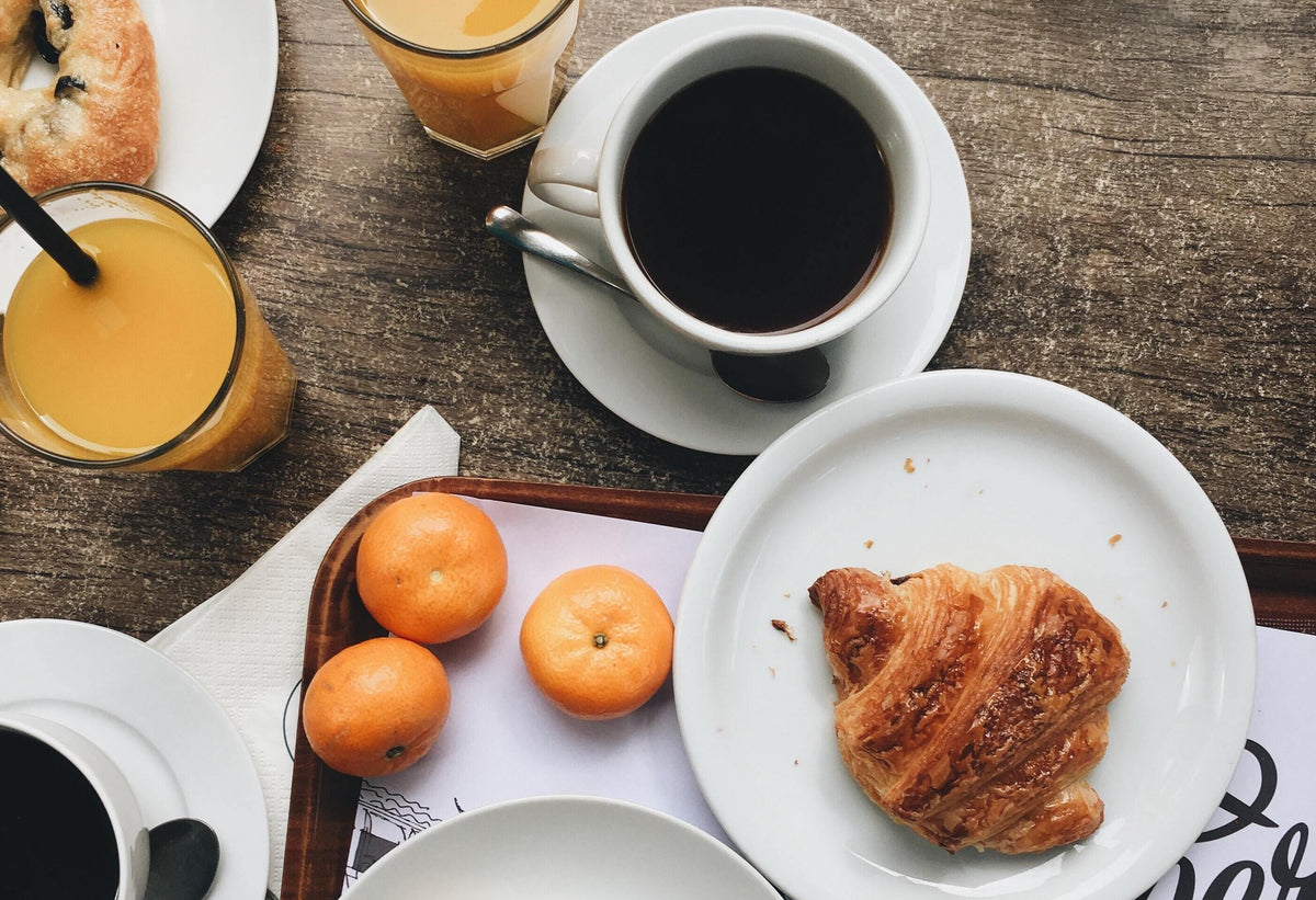 Overhead view of a breakfast spread with a cup of black coffee, glasses of orange juice, a croissant, and tangerines on a wooden table