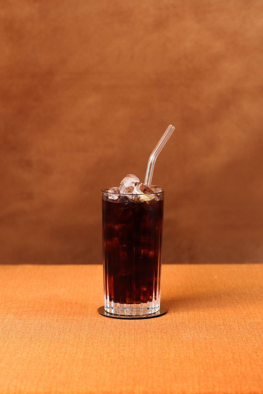 Front view of Classic Cold Brew in a tall ribbed glass with ice and a clear glass straw on an orange table and warm brown backdrop