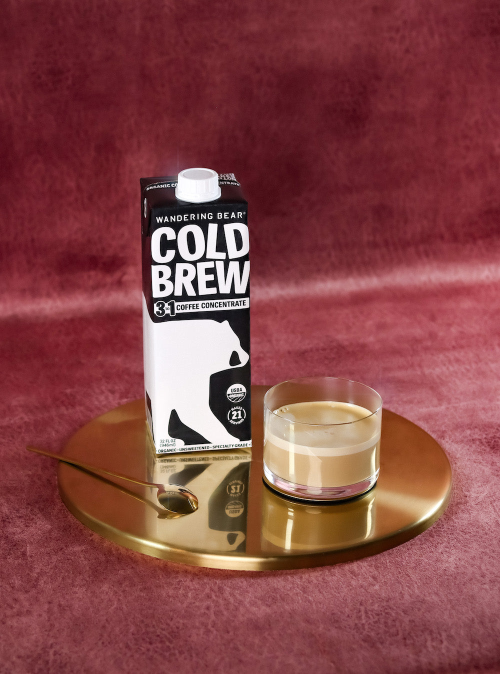 Wandering Bear 3 to 1 Cold Brew carton with label reading COLD BREW beside a cortado glass on a gold tray against a burgundy velvet background