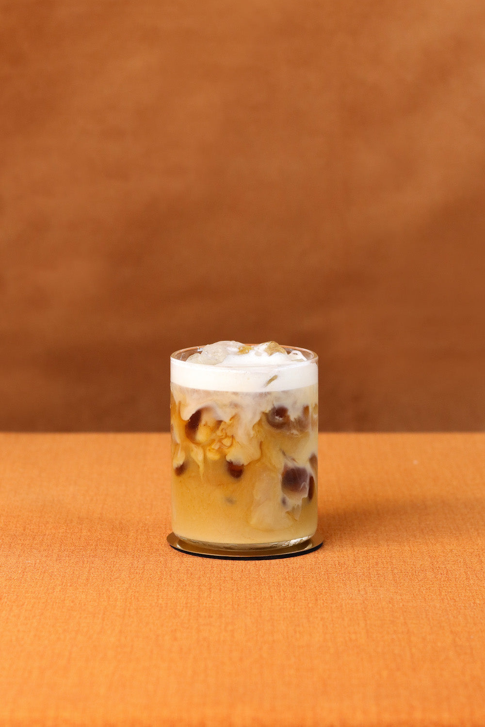 Front view of iced cold brew cappuccino showcasing milk swirl, ice, and foam in a clear glass on an orange fabric background