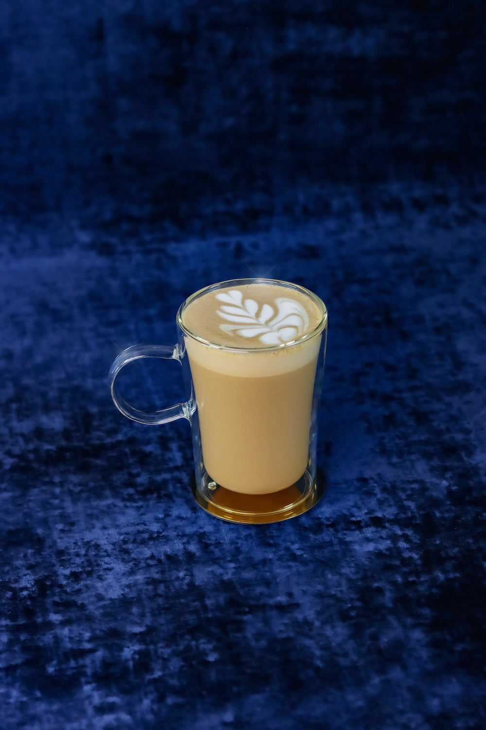 Front view of cold brew latte in a clear double-walled glass mug with leaf latte art on a deep blue velvet background