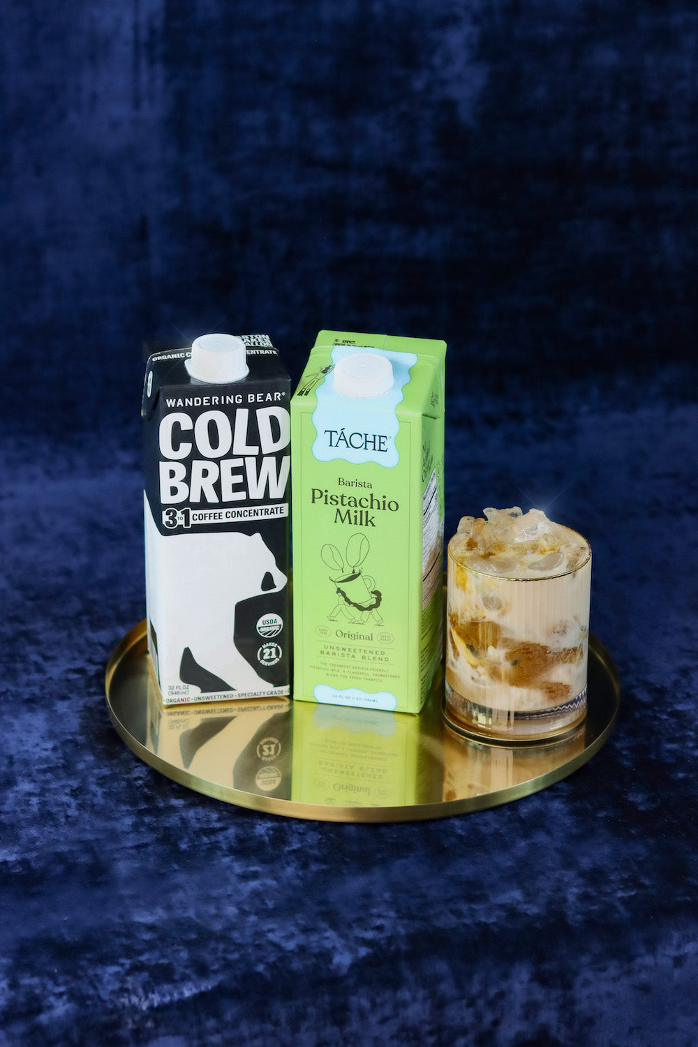 Iced pistachio latte beside Wandering Bear Cold Brew concentrate 3:1 and Tâche pistachio milk cartons on gold tray with blue backdrop