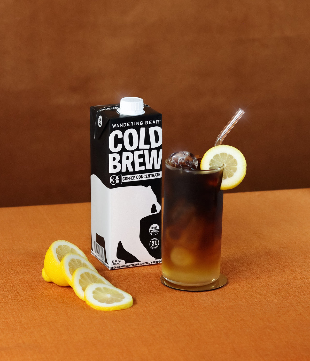 Glass of iced Arnold Palmer with lemon slice next to Wandering Bear carton labeled COLD BREW on orange background