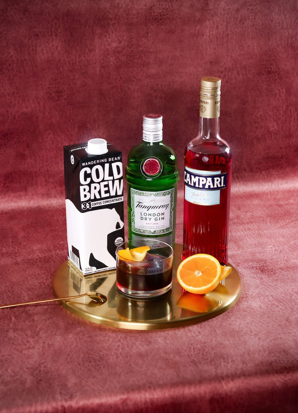 Negroni cocktail posed with Wandering Bear Cold Brew cold brew concentrate 3:1 carton, Tanqueray gin bottle, and Campari on a gold tray with orange garnish and red velvet background
