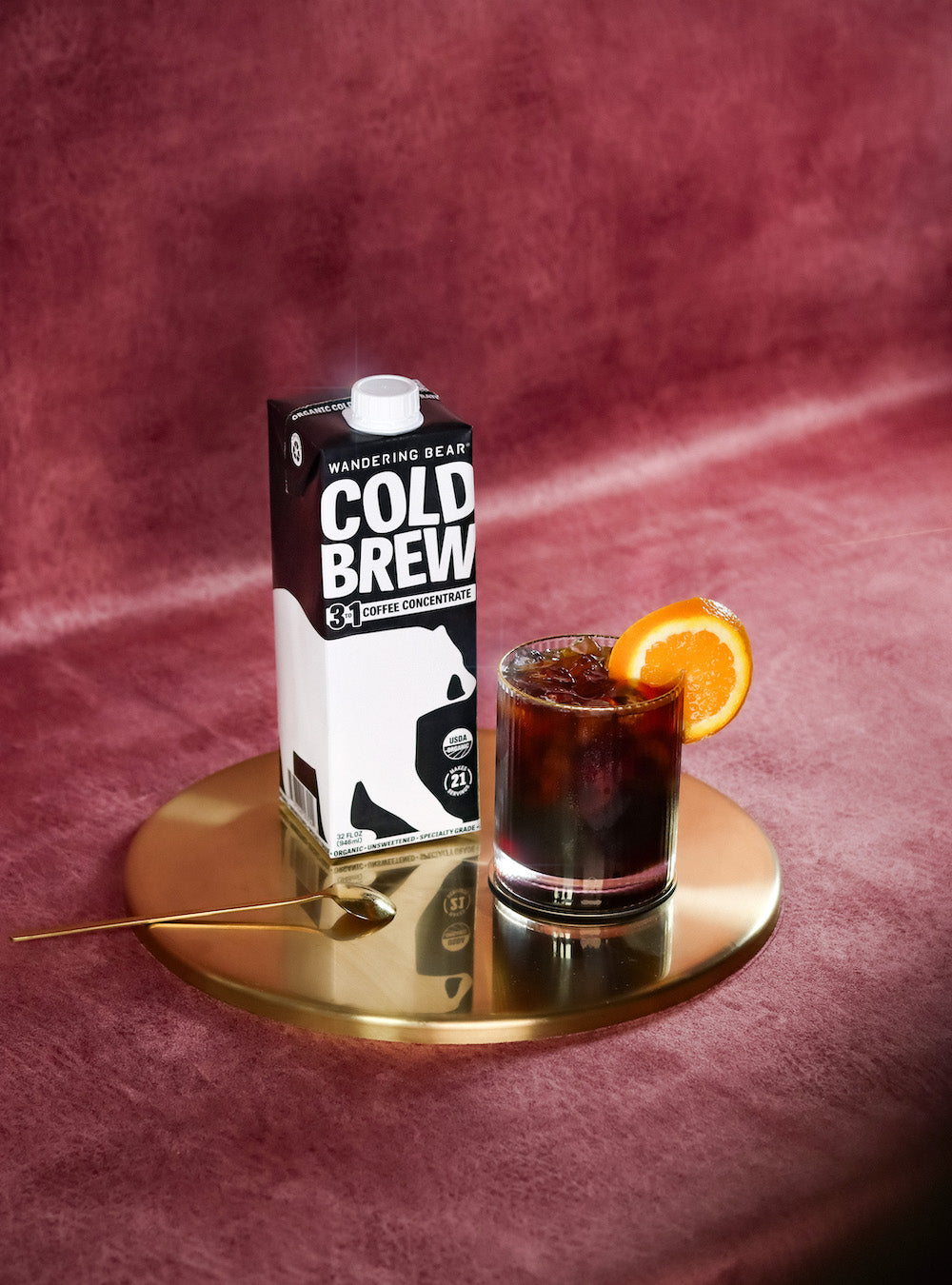 Glass of sparkling cold brew soda garnished with an orange slice beside a Wandering Bear Cold Brew carton on a gold tray against a pink velvet background