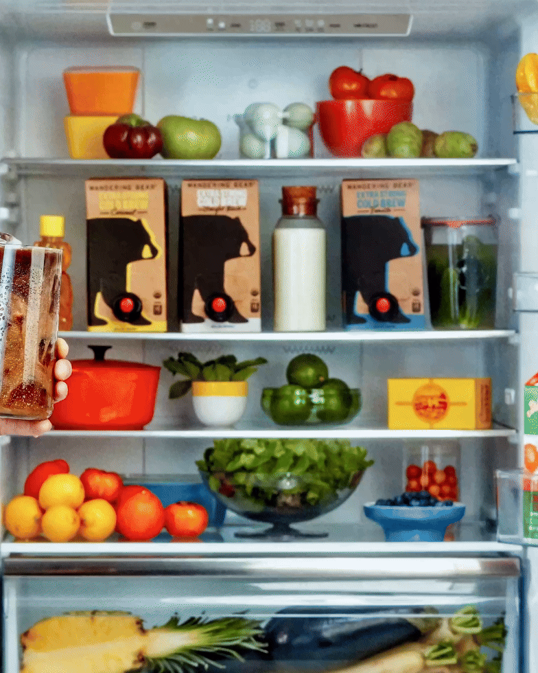 Hand holding iced coffee glass in front of an open fridge with Wandering Bear cold brew boxes, milk, and fruit