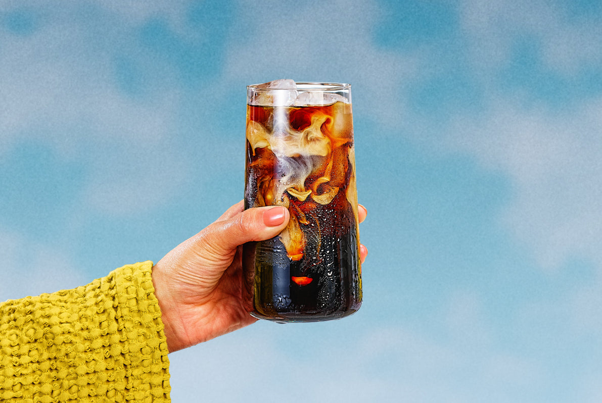 Hand holding a tall glass of iced black coffee with cream swirling against a soft blue sky background