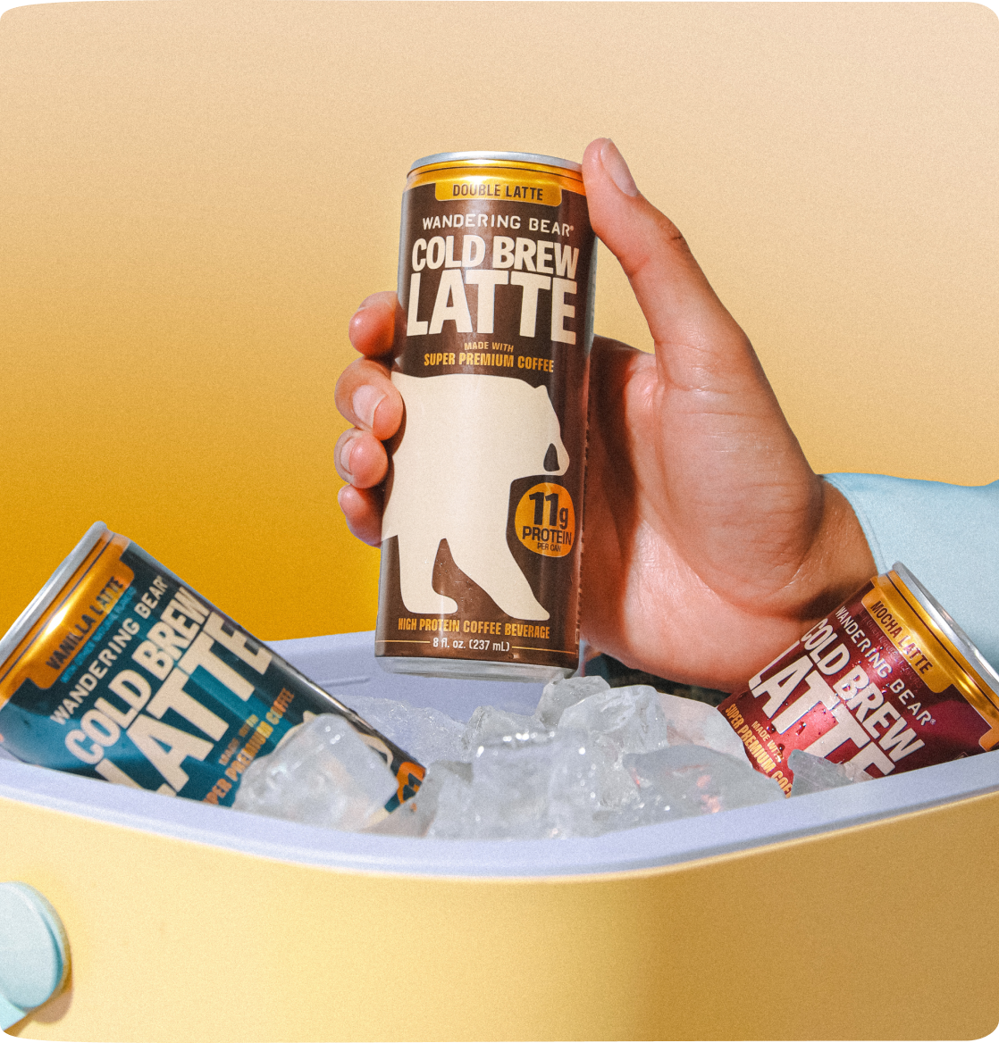 Hand-held front view of 8oz Cold Brew Latte can showcasing bear logo and 11g protein badge with two cans chilled in an ice-filled yellow cooler